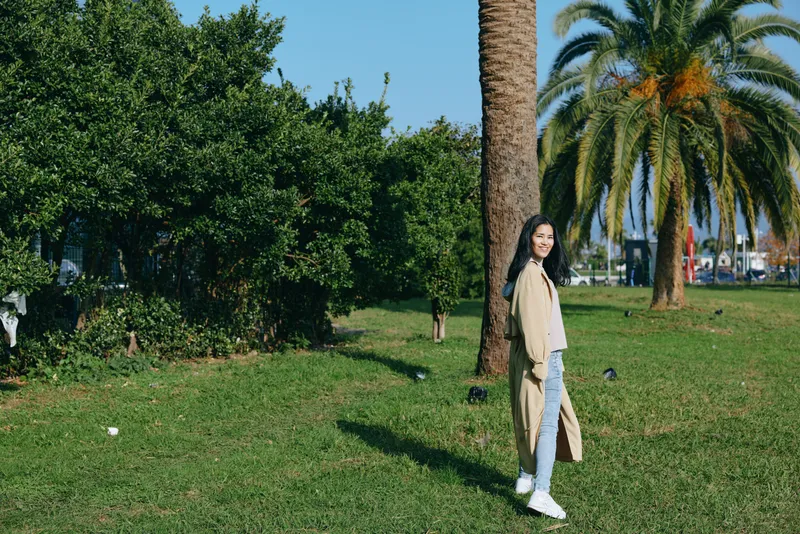 Woman strolling through a sunny park with palm trees, green grass and open space, wearing a beige coat and white sneakers, conveying relaxed outdoors lifestyle, nature, and casual fashion in daylight.