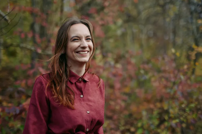 Portrait of a smiling woman outdoors in autumn, wearing a burgundy shirt. The image emphasizes authenticity and honest emotion with natural light and relaxed posture.