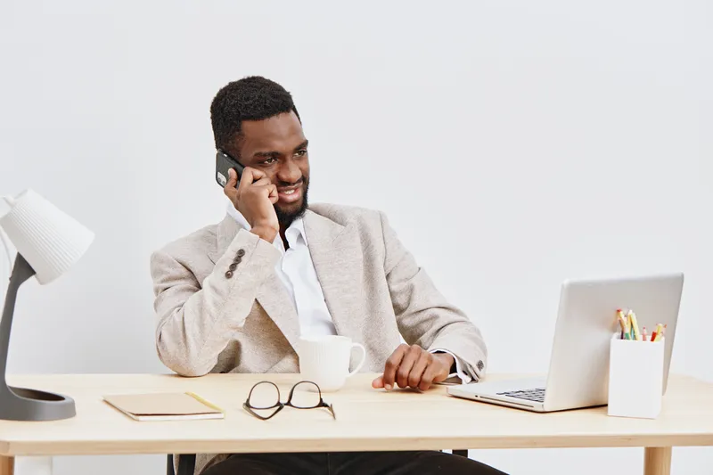 Professional smiling man working at desk talking on mobile phone in modern office environment with laptop, documents, and glasses on table, embodying business communication and productivity, studio sh