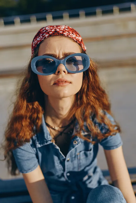 lifestyle woman wearing oversized blue sunglasses and red patterned headband in modern boho-western denim street style with film color effect and natural sunlight