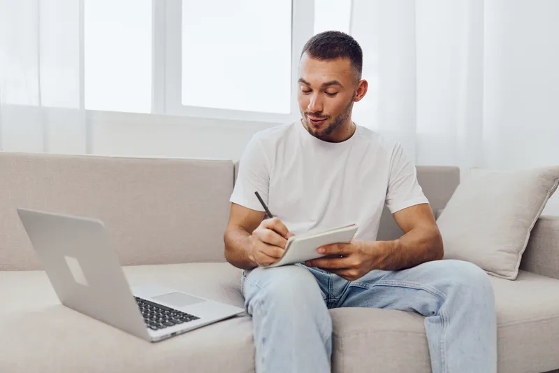 Thoughtful man sitting on a sofa, writing in a notebook, surrounded by a cozy interior, showcasing creativity and productivity in a modern lifestyle.