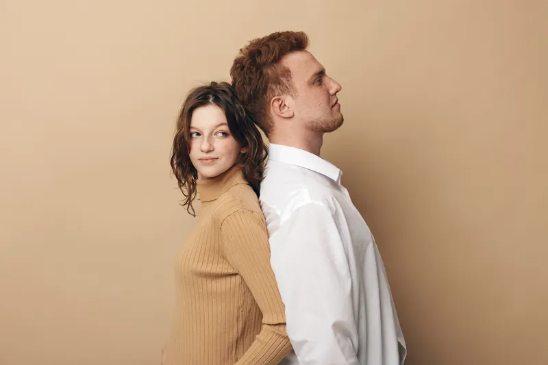 Young man and woman stand back to back against beige background. Casual clothing, relaxed mood, diverse couple with natural expressions. Concept of relationship, friendship, and connection.
