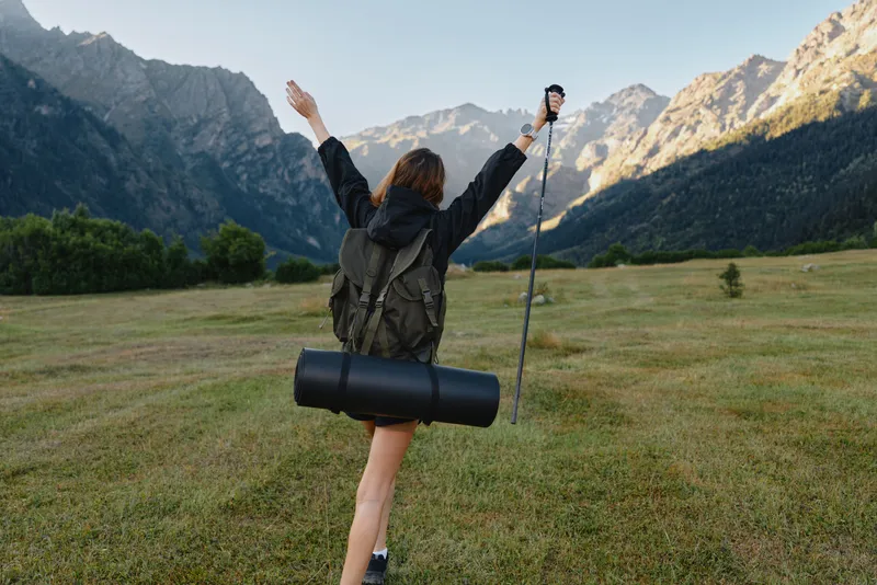 A joyful hiker stands in a wide meadow beneath towering mountains, arms raised in triumph. She carries a backpack and gear, embracing the wild outdoor journey.