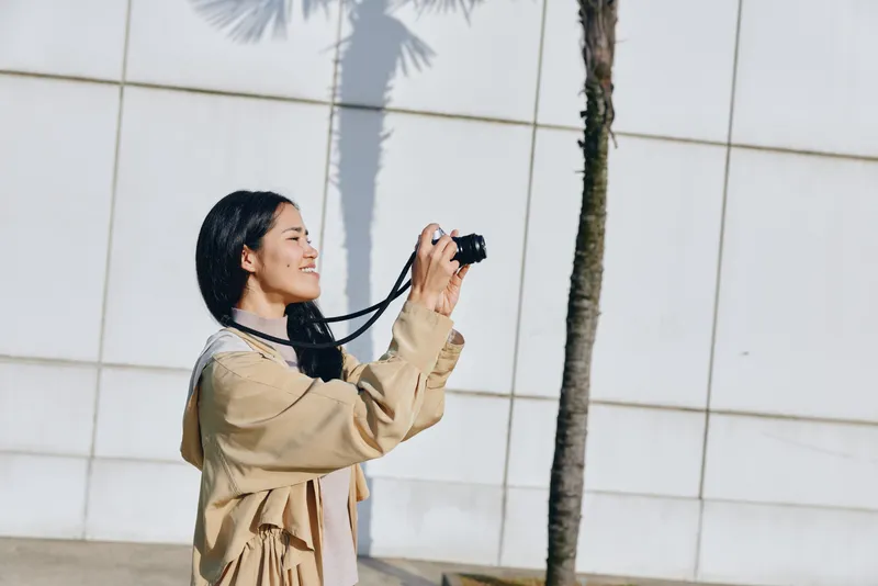 Woman with camera in an outdoor photography scene beside a palm and a modern wall, casual attire, smiling as she shoots on a sunny day, capturing travel vibe with street style.