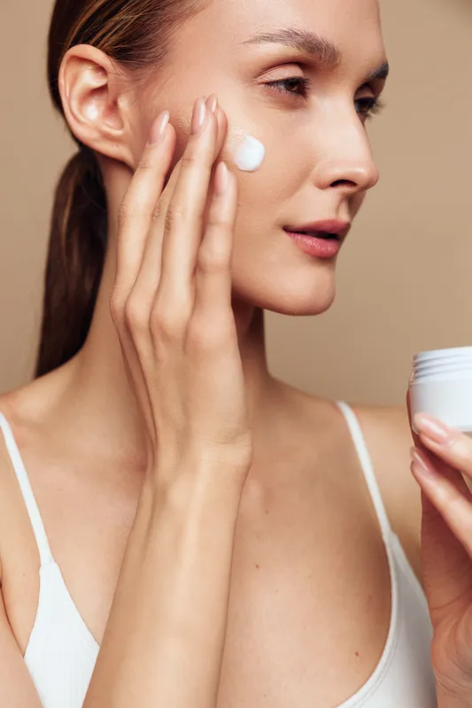A young woman with clear skin applies white cream on her cheek using fingers. She holds an open jar in her other hand against a neutral background. Concept for skincare and beauty routine.