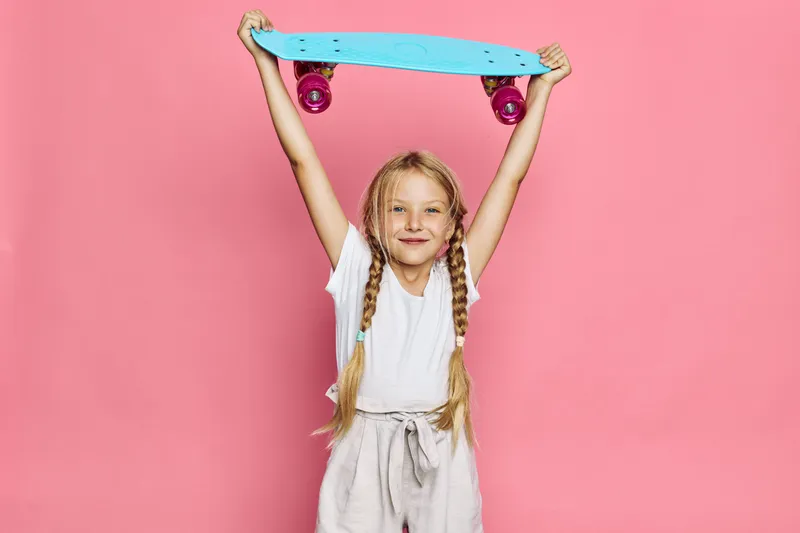 Smiling girl with braided hair holds a blue skateboard above her head on a pink background. Casual clothes and confident expression. Sport and childhood concept.
