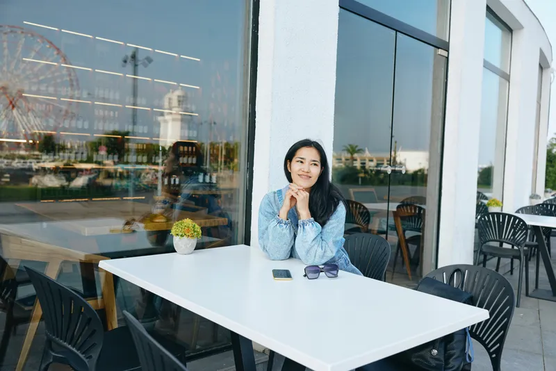 Woman sits at a white outdoor cafe table with thoughtful expression, glass facade reflections, sunny day, small plant, sunglasses on table, casual denim shirt, relaxed urban cafe vibe