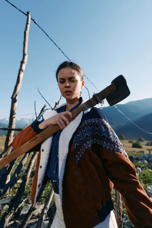 Woman with axe in countryside mountains, portrait by wooden fence outdoors. Young woodcutter in patterned jacket holding tool, rural lifestyle and rugged nature scene and solitude.