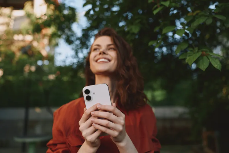Cheerful woman outdoors in a park, lifestyle portrait, smiling while holding a smartphone, casual red top, sunlit leaves, candid moment of connection and happiness, social interaction