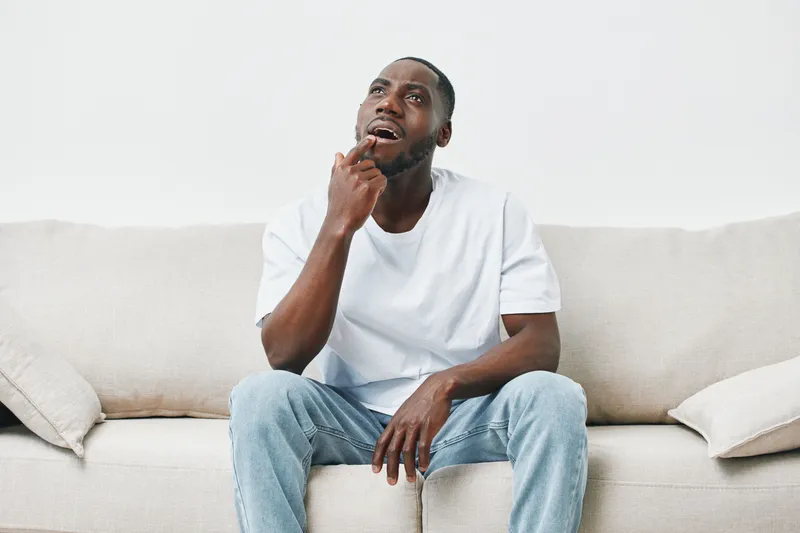 Thoughtful young Black man in casual attire sitting on a couch, reflecting with an expressive gesture, evoking emotions of introspection and contemplation
