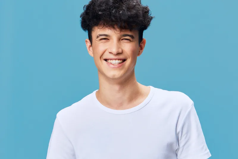 Smiling man with curly hair wearing a white t-shirt stands against blue background, expressing happiness and positive energy with bright casual style.