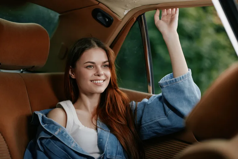 Young woman with long hair smiling in a vintage car, wearing a denim jacket, surrounded by greenery. Natural light enhances the cheerful mood.
