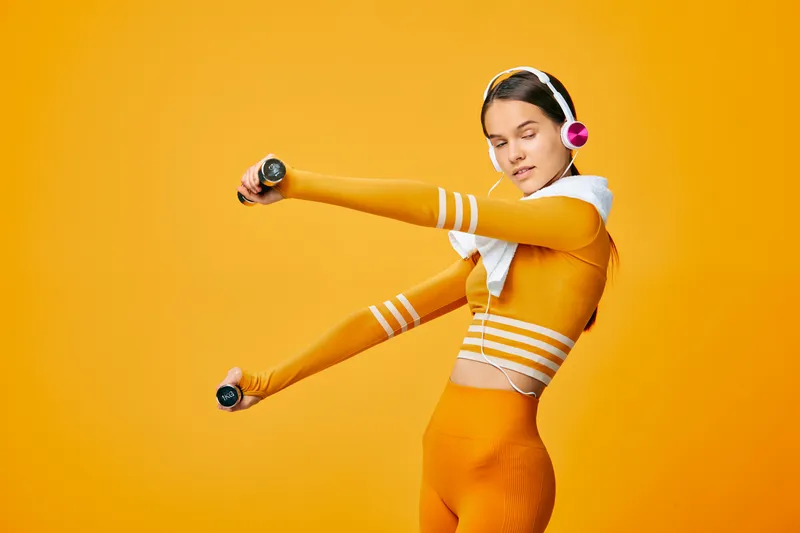Fitness woman in orange athletic wear with headphones and towel, holding dumbbells and performing a dynamic stretch in a bold orange studio.