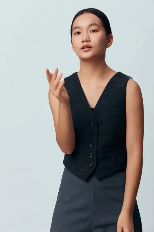 Young woman with long black hair wearing dark vest and skirt looks confidently with natural makeup and relaxed pose on plain light background in studio portrait.