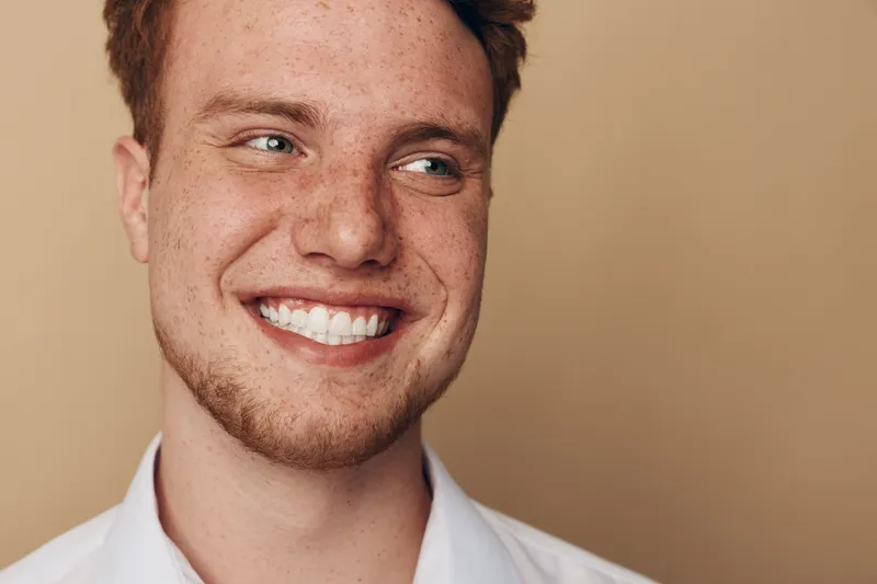 Smiling young man with red hair and freckles looks away, showing white teeth and joyful expression. Casual portrait on beige background with natural lighting and positive mood.