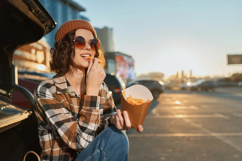 Woman eating fries by a car trunk at golden hour, wearing sunglasses, beanie and plaid coat, smile and relaxed vibe. Candid lifestyle portrait with authenticity, golden hour glow.