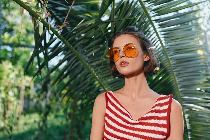 A stylish young woman stands outdoors among lush tropical foliage wearing a red and white striped sleeveless top and round orange sunglasses, exuding a chic summer mood.