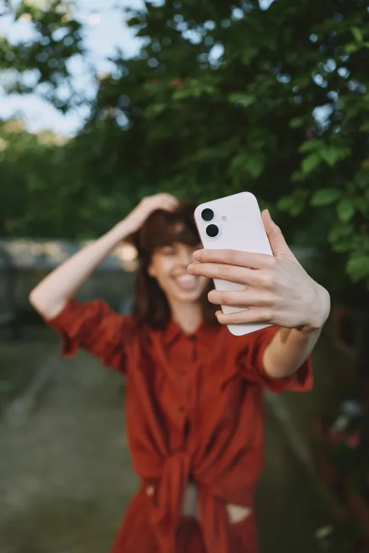Vibrant woman lifestyle scene outdoors, smiling while taking a selfie in bright sunlight, casual red dress, lush greenery background, candid cheerful moment