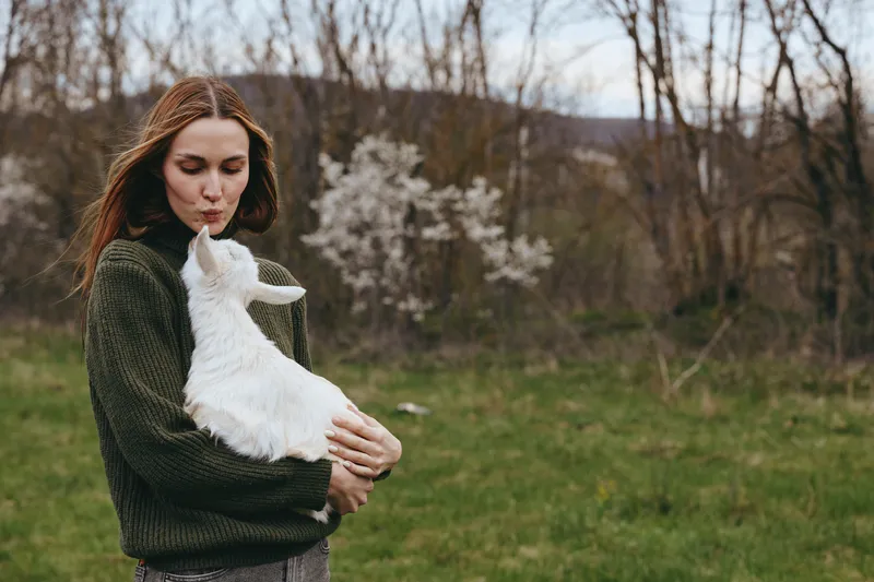 Calm outdoor portrait of a woman in a green knit sweater tenderly holding a white goat kid in a grassy field, showcasing loving countryside lifestyle and connection with animals under soft natural