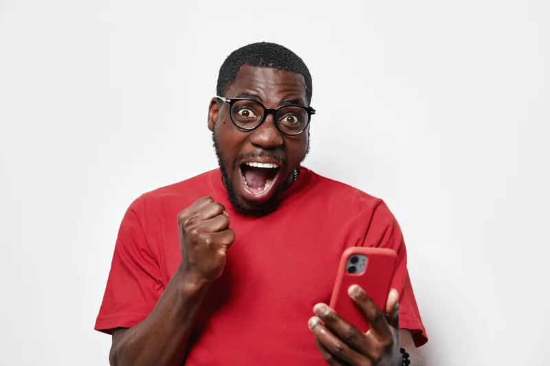 Excited African American man with dark skin wearing glasses and red shirt holding smartphone and celebrating success with clenched fist on white background in studio.