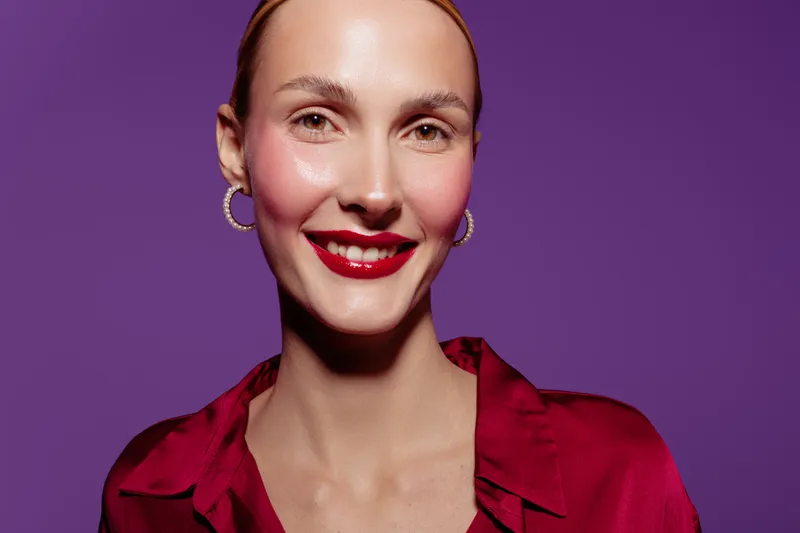 Smiling woman with red lipstick and hoop earrings wears a red satin blouse against purple background. Portrait of confident female model with natural skin and bright makeup in studio lighting.