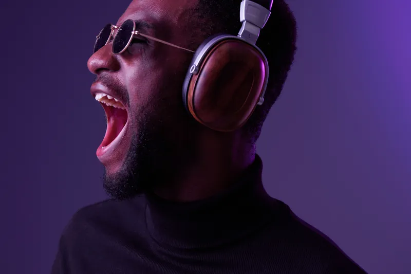 Young African man shouting joyfully while wearing stylish headphones and sunglasses against a vibrant purple background, expressing passion for music
