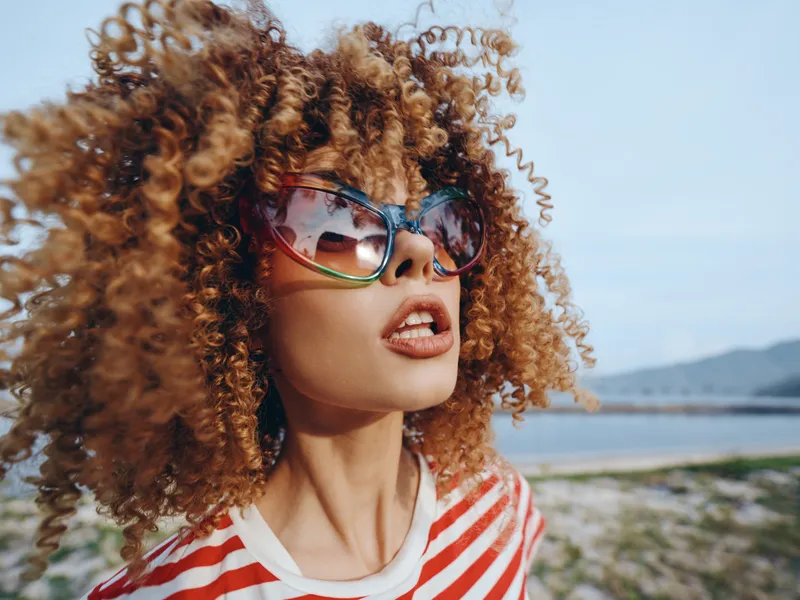 Portrait of a stylish woman with voluminous curly hair and oversized sunglasses at the beach, wind tousling locks against a bright seaside backdrop.