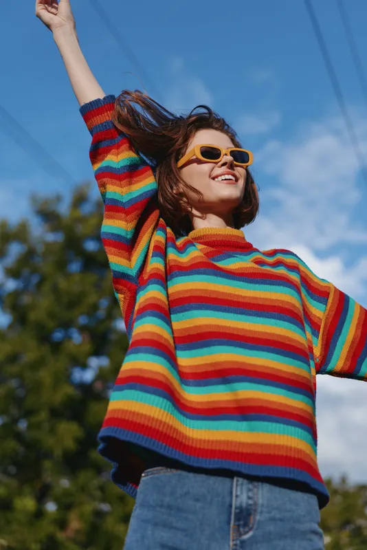 Young woman in a rainbow sweater and yellow sunglasses smiling outdoors with raised arms, casual jeans and curly hair, carefree sunny lifestyle portrait for travel and fashion.