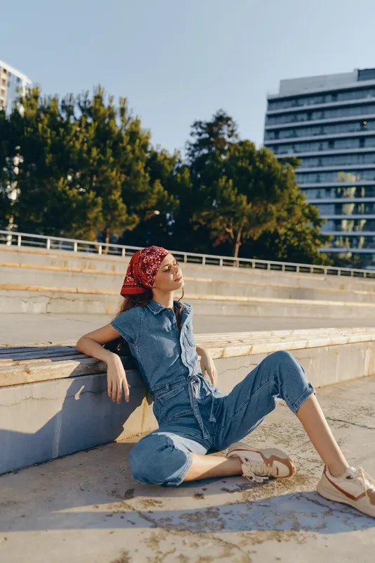 lifestyle woman wearing modern boho-western denim street style outfit with red bandana relaxing outdoors in warm sunlight, casual summer fashion, film color atmosphere