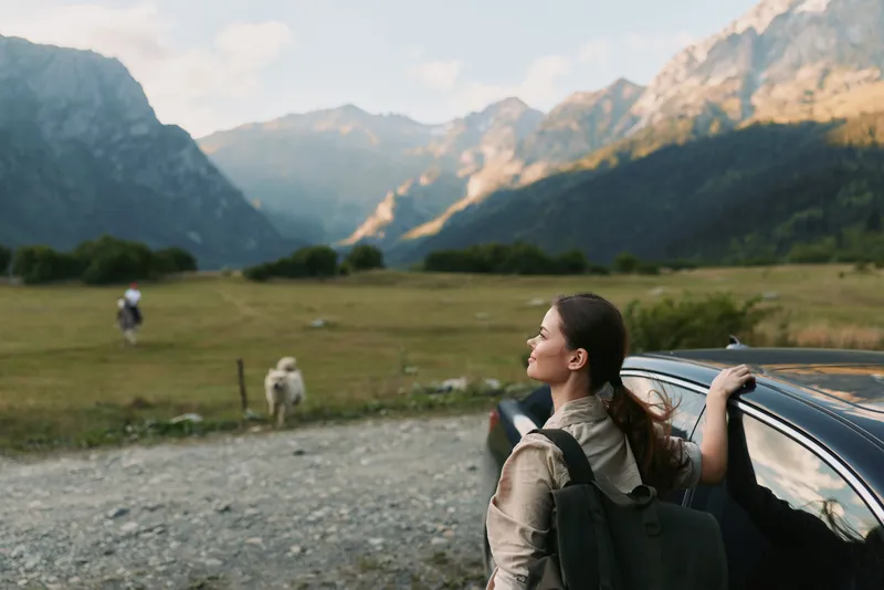 Cheerful woman outdoors beside a car with backpack, mountains in the distance, sunlit meadow, calm lifestyle moment, clear sky, natural colors, breeze