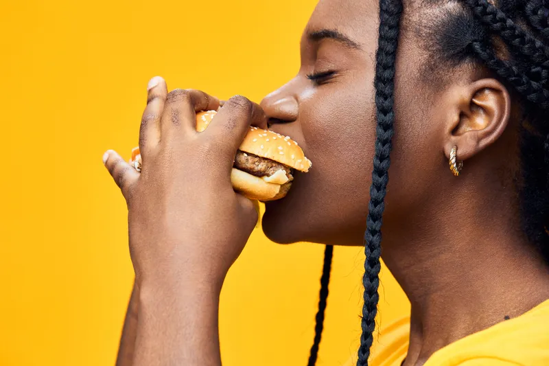 Young woman enjoying a delicious burger against a vibrant yellow background, showcasing her love for fast food and indulgence
