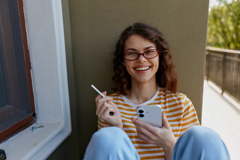 Smiling young woman with glasses sits on a doorway inside a home, holding a smartphone and a cigarette, wearing a yellow striped shirt and light blue jeans, casual relaxed mood, she leans against a