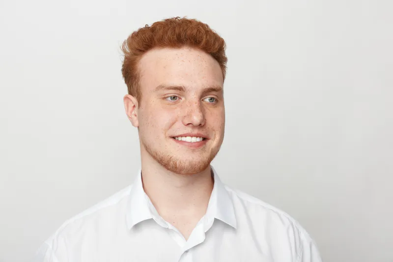 Smiling young man with red hair and freckles wearing white shirt looks away on white background. Portrait of confident male model with casual style and natural expression in studio.