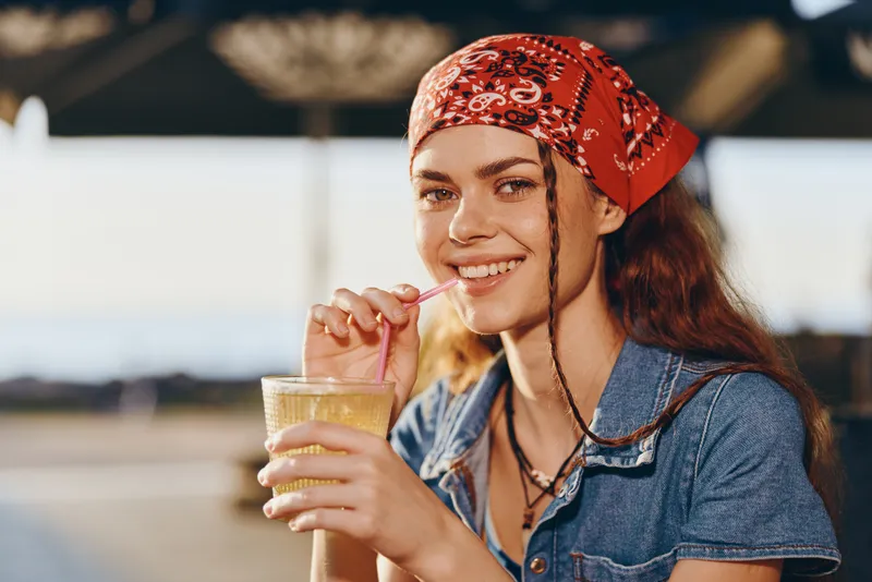 smiling woman with red bandana drinking refreshing juice outdoors on sunny day wearing denim shirt and casual necklace