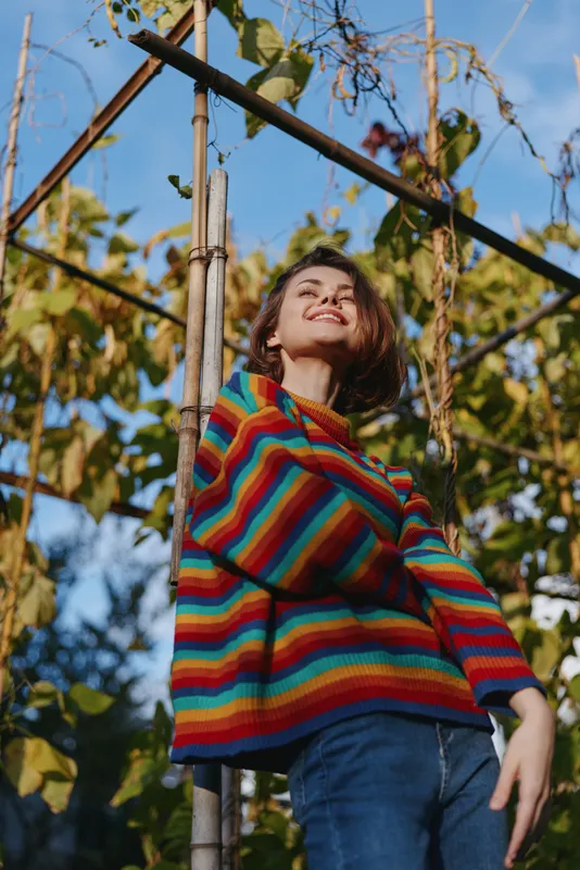 Woman smiling in rainbow sweater leaning on bamboo trellis in garden, outdoor portrait with sunlight and blue sky, casual colorful fashion, joyful expression and relaxed pose.