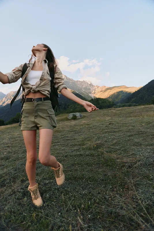 Joyful woman in outdoor lifestyle, wearing beige jacket and shorts, dances across grassy hillside with mountains in the background, expressing freedom and adventure.