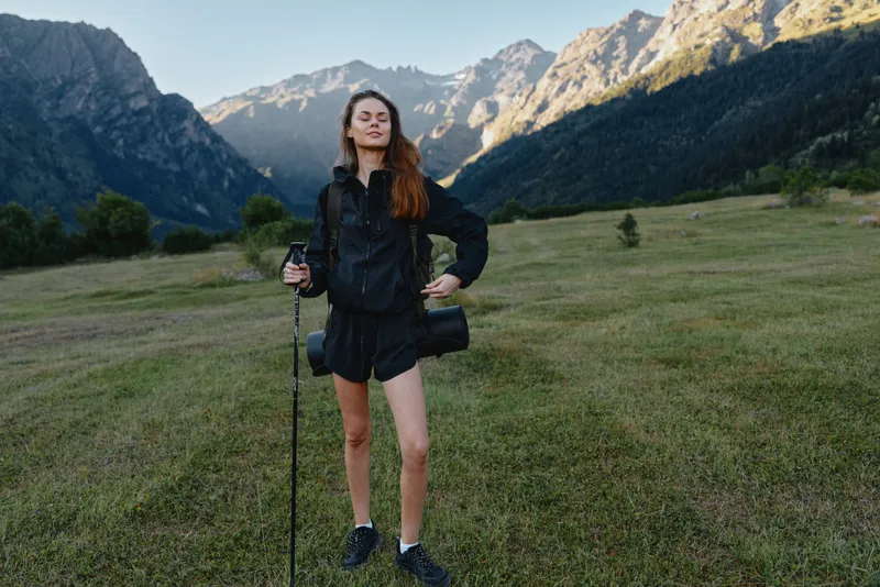 A confident hiker stands in a wide mountain meadow, holding a trekking pole with a small bag. She gazes ahead, serene and adventurous, ready to explore nature.