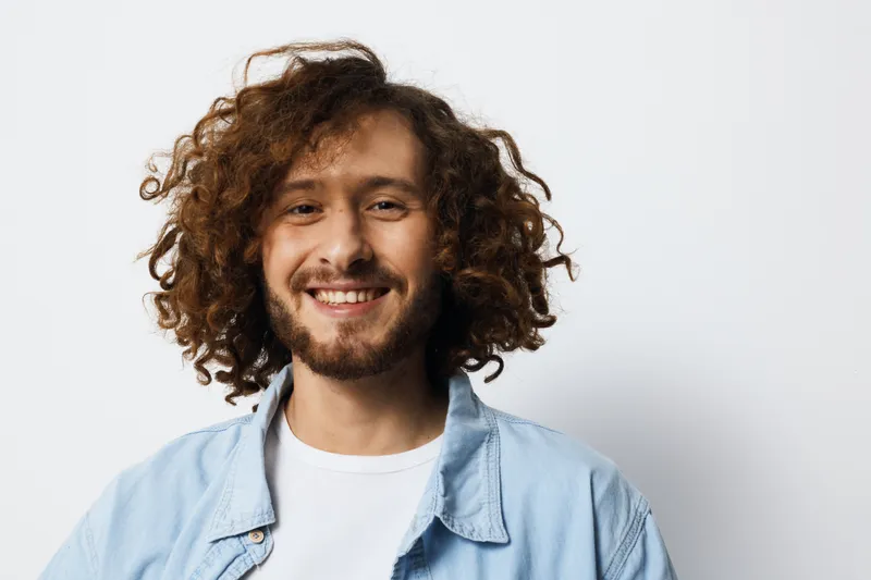 Cheerful young man with curly hair smiling against a clean background, dressed casually in a light blue shirt, embodying a positive and friendly demeanor