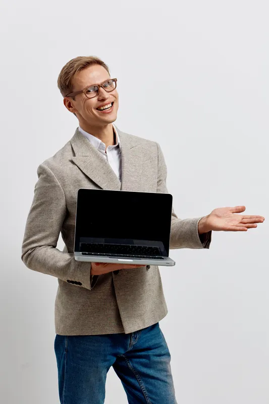 Smiling man holding a laptop in one hand and gesturing with the other, dressed in beige blazer and casual jeans, posing against a plain white background. Studio portrait for business, technology, and 