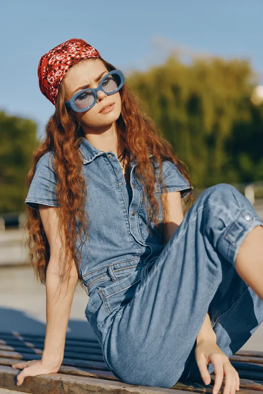 lifestyle woman in modern boho-western denim street style sitting outdoors with long red hair, blue sunglasses, and bandana, captured in warm film color tones