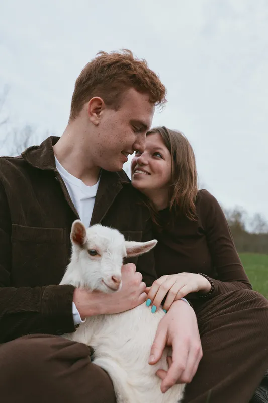 A couple sits outdoors with a young goat kid, showing affection and smiles, creating a warm lifestyle scene in a rural setting with a green field and soft light, capturing tenderness and connection.
