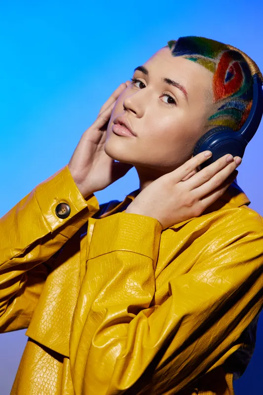 Creative woman with colorful hair wearing headphones and a yellow leather jacket, posing against a vibrant blue background with a thoughtful expression.