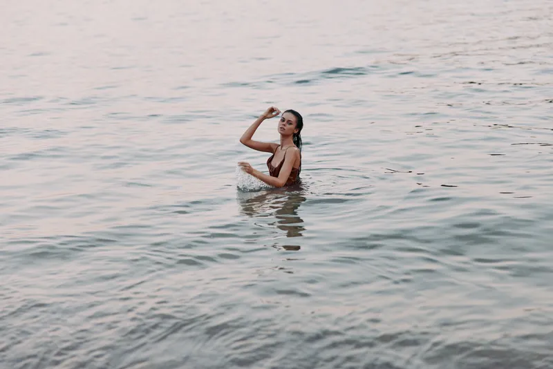 A lone swimmer stands waist deep in calm water, gazing to the side with a relaxed pose as soft evening light highlights gentle ripples around her silhouette.