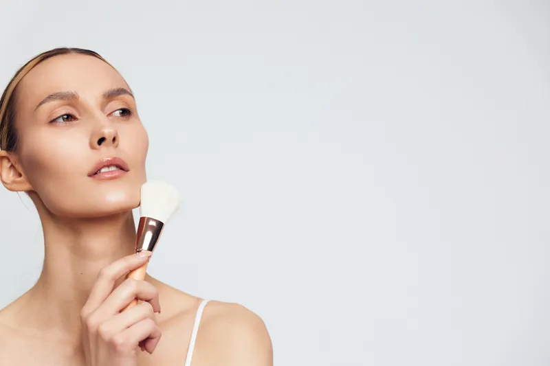 Young woman with clear skin applying powder makeup with a brush on her face. Minimalist background and soft lighting emphasize beauty and skincare routine.