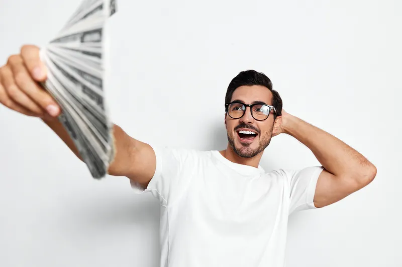 Happy cheerful young man wearing glasses holds a stack of money while smiling at the camera, isolated on a plain white background. Lifestyle finance concept.
