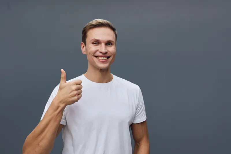 Cheerful happy man in white t-shirt showing thumbs up gesture smiling at camera isolated on gray background studio portrait. People lifestyle concept