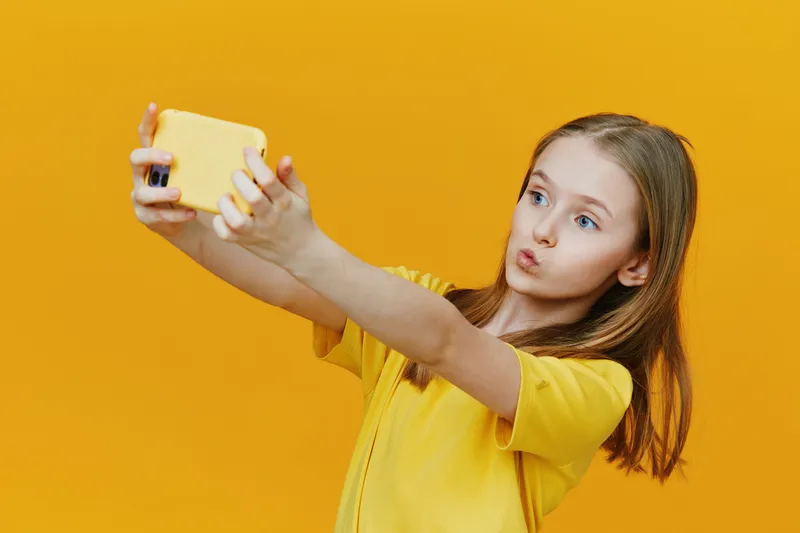 A young girl with straight hair wearing a yellow t shirt takes a selfie against a bright yellow background, showcasing joy and playfulness in her expression
