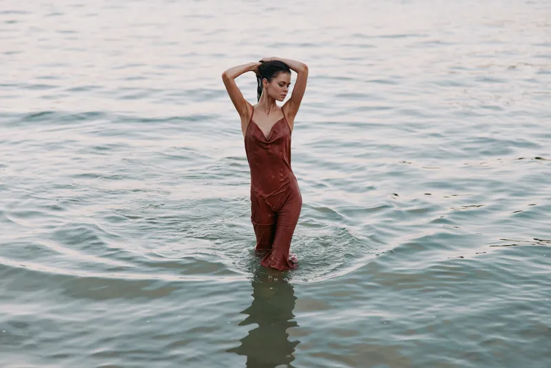 A woman in a burgundy dress stands waist-deep in a calm sea, striking a graceful pose with arms lifted, creating a bold fashion moment and shimmering water reflections.