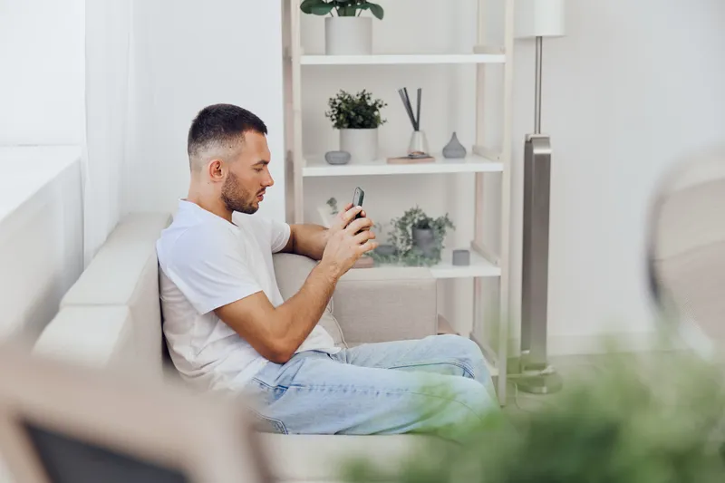 Young man sitting on a couch, focused on his smartphone, surrounded by houseplants in a bright, modern living room, creating a sense of contemporary lifestyle.