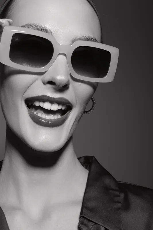 Close-up of a happy caucasian woman with fair skin wearing large sunglasses and hoop earrings smiling with open mouth. Concept of fashion, joy, and beauty in studio lighting.